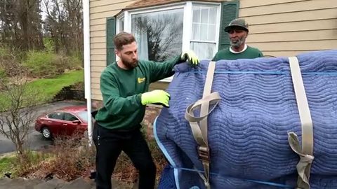 two movers securing straps on a blanket-wrapped upright piano on a board, face visible